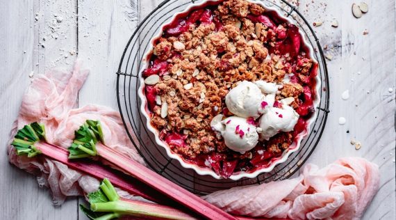 Eine Tarteform mit Rhabarbercrumble von oben fotografiert, darauf drei Kugeln Vanilleeis, daneben, Rhabarberstangen