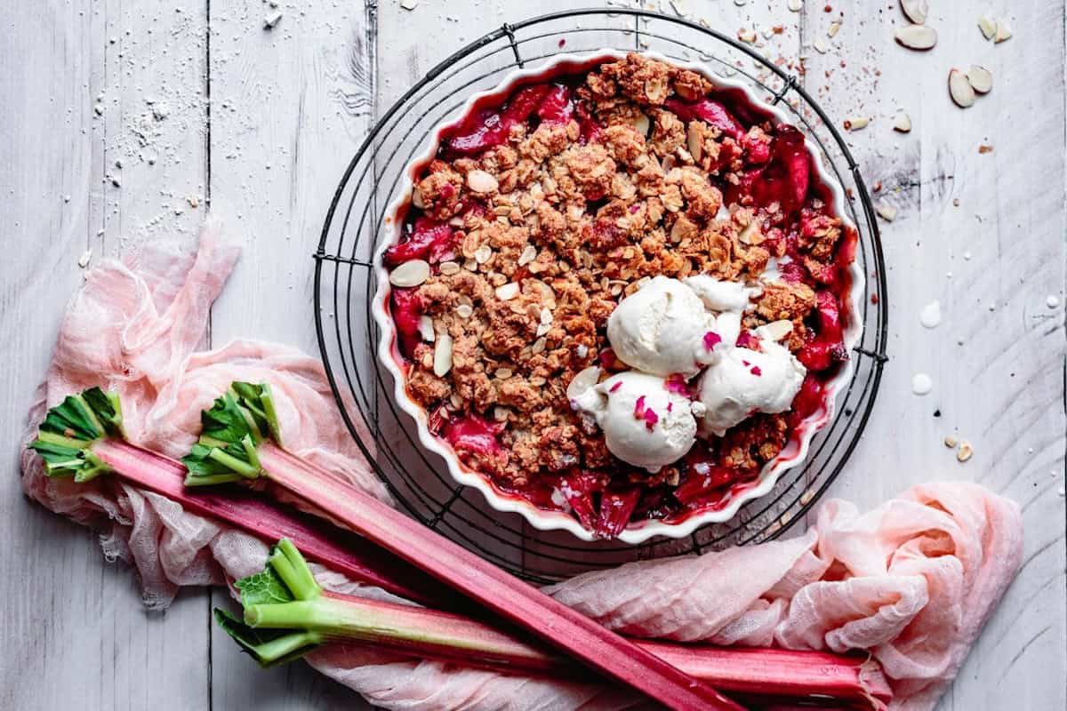 Eine Tarteform mit Rhabarber-Crumble von oben fotografiert, darauf drei Kugeln Vanilleeis, daneben, Rhabarberstangen.