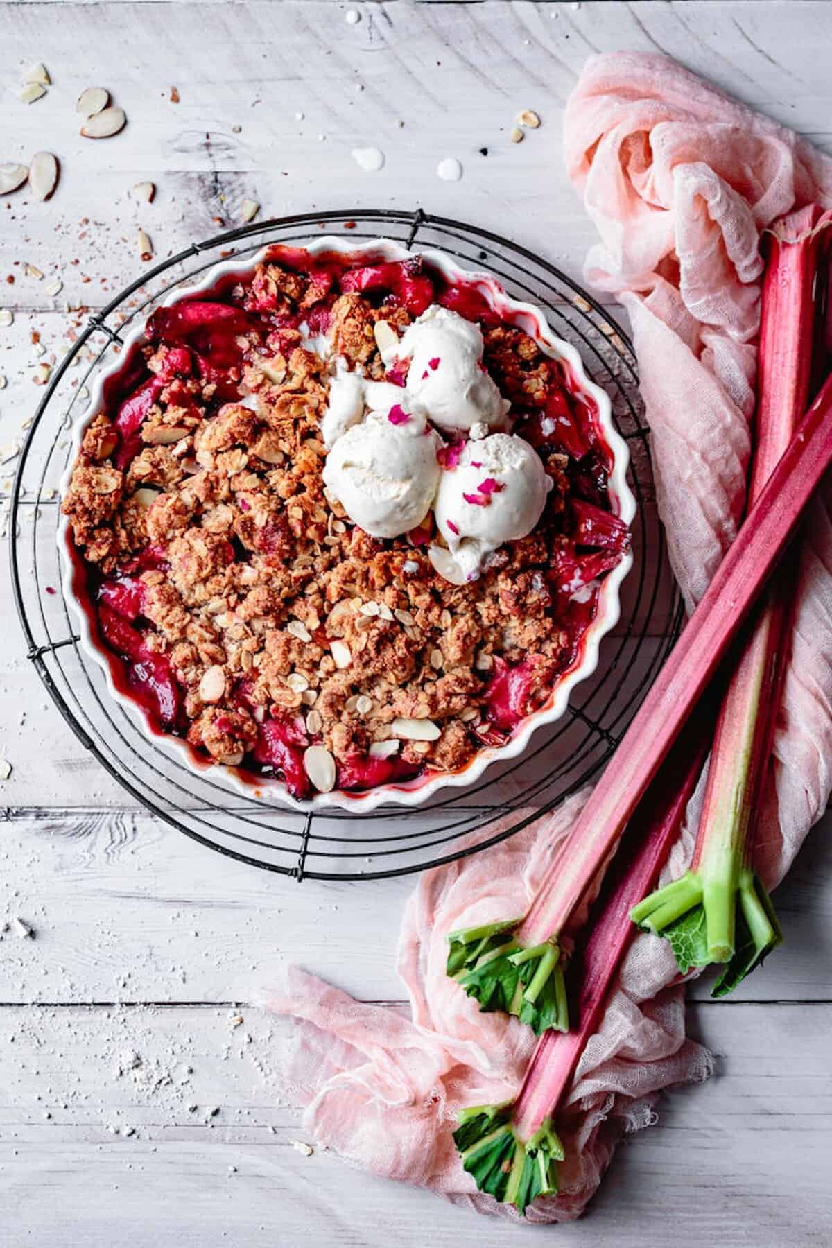 Eine Tarteform mit Rhabarbercrumble von oben fotografiert, darauf drei Kugeln Vanilleeis, daneben, Rhabarberstangen