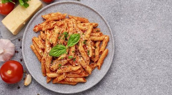 Penne All'arrabbiata auf einem grauen Teller von oben fotografiert, danebn Tomaten, Kräuter und Käse