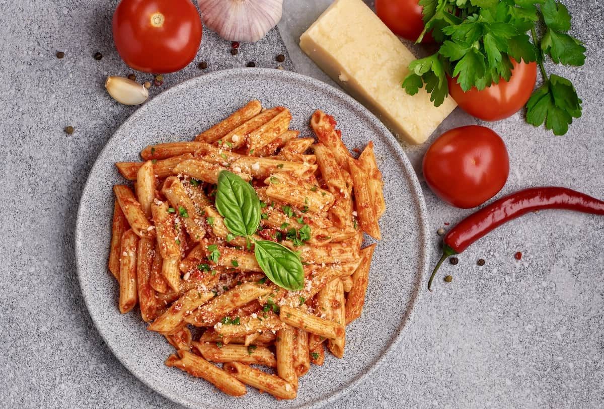 Penne All'arrabbiata auf einem grauen Teller von oben fotografiert. Daneben Basilikum, Tomaten, Chili, Käse