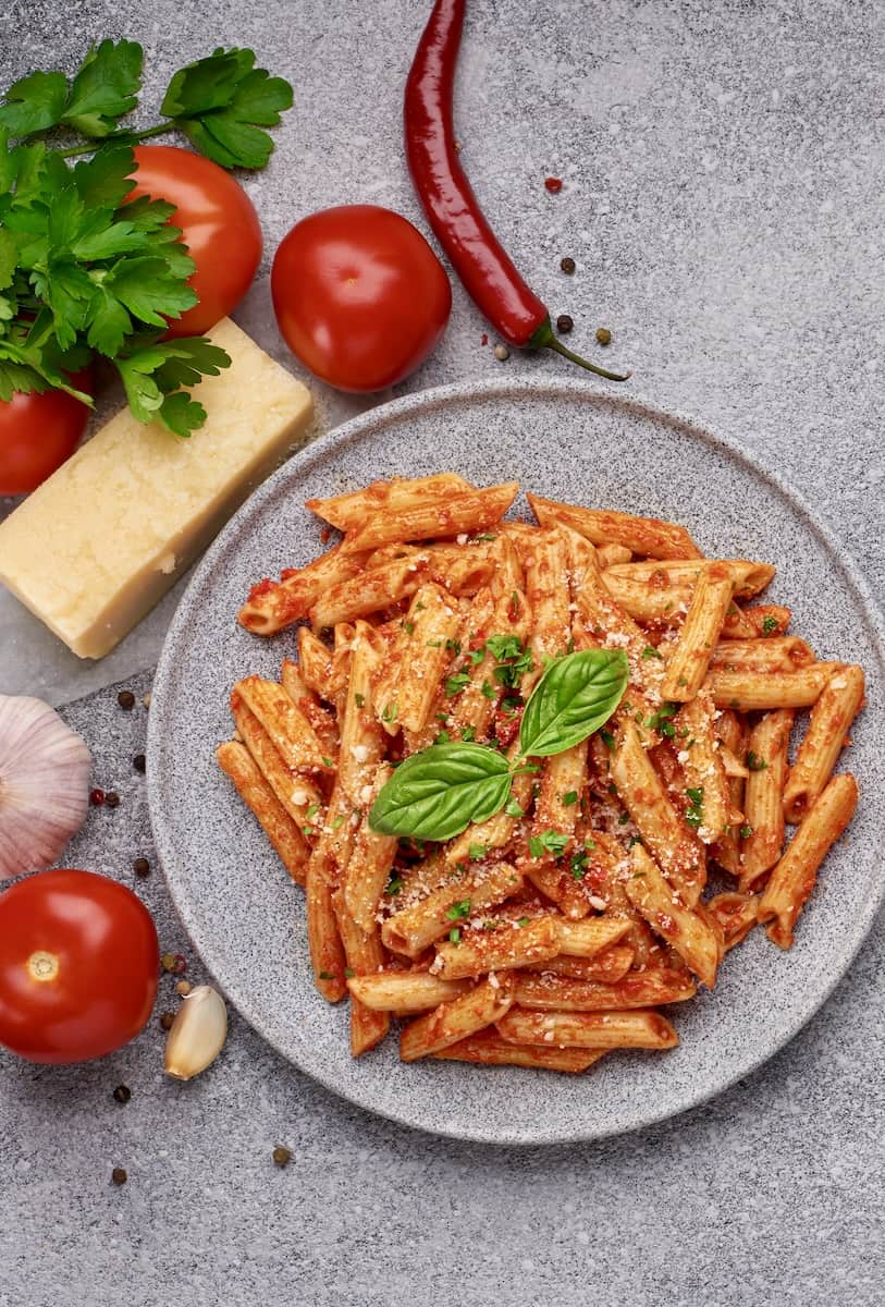 Penne All'arrabbiata auf einem grauen Teller von oben fotografiert. Daneben Basilikum, Tomaten, Chili, Käse