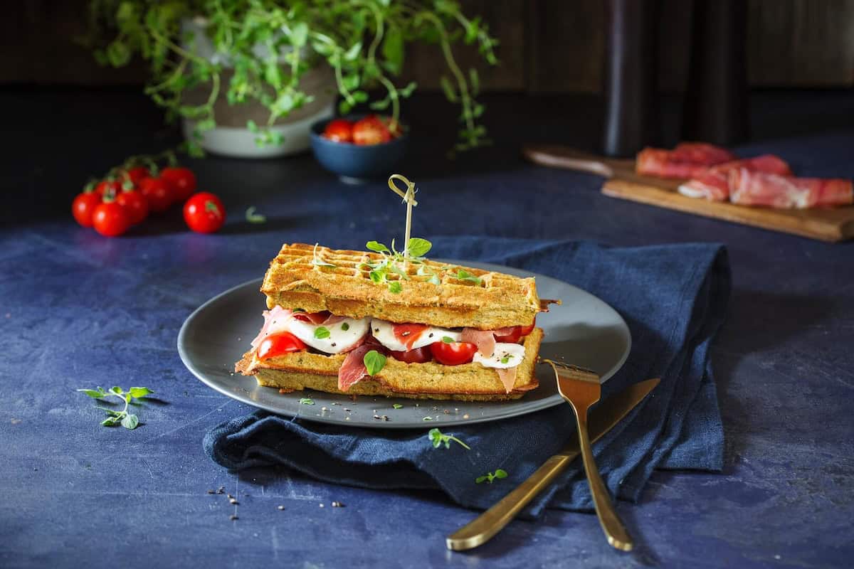 Ei Sandwich aus Vollkorn-Kräuterwaffeln mit Tomate-Mozzarella auf grauem Teller und blauem Untergrund – Foto: Anna Gieseler