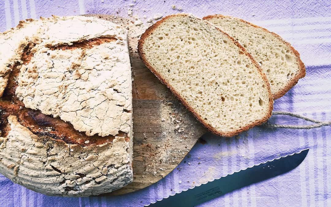 Ein rundes Brot liegt auf einem Brett und wurde aufgeschnitten. Zwei Scheiben davon, sowie das Brotmesser liegen daneben.