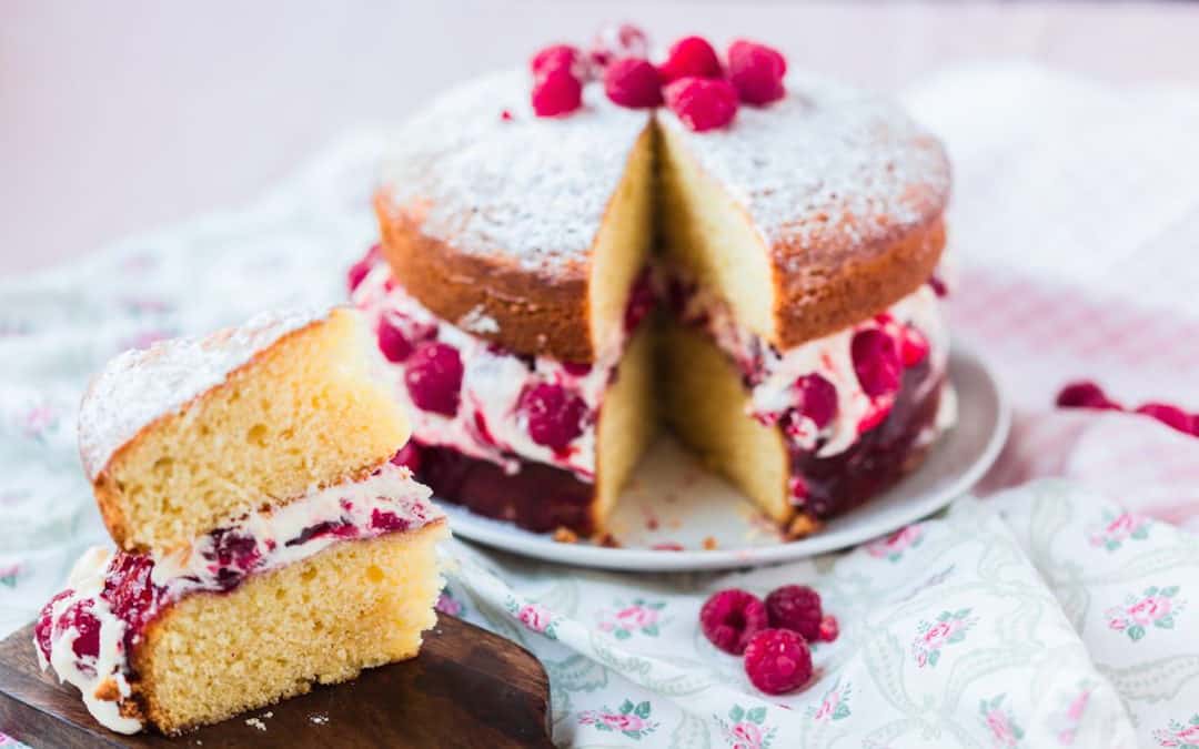 Ein Himbeerbiskuitkuchen liegt angeschnitten auf einem Teller. Im Vordergrund ist das abgeschnittene Stück mit der Himbeerschicht zu sehen.