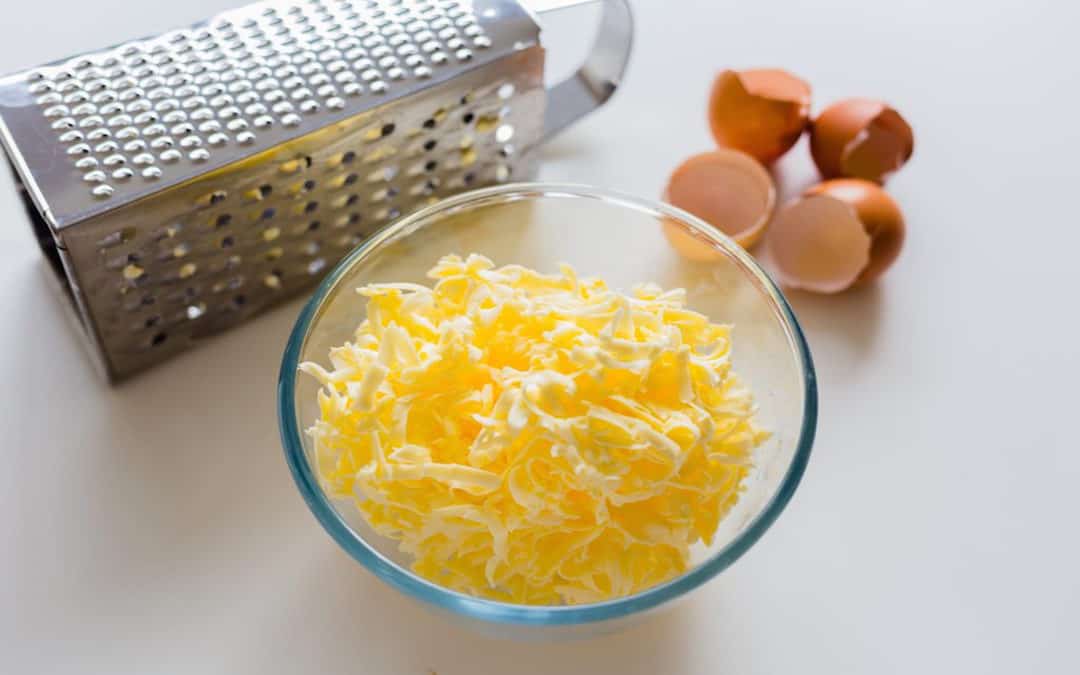 Butter steht in geraspelter Form in einer Glasschüssel vor ein paar Eierschalen und einer Raspel.