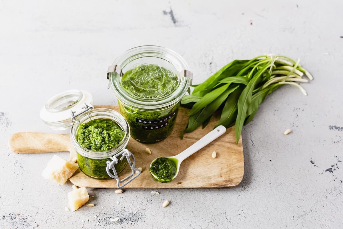 Zwei Gläser Bärlauch-Pesto auf einem Holzbrett, daneben frischer Bärlauch