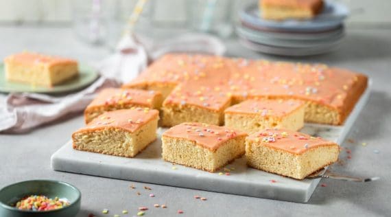 Flacher Blechkuchen mit Zuckerguss in Orange und Streuseln, mehrere Stücek auf einem Teller – Foto: Anna Gieseler