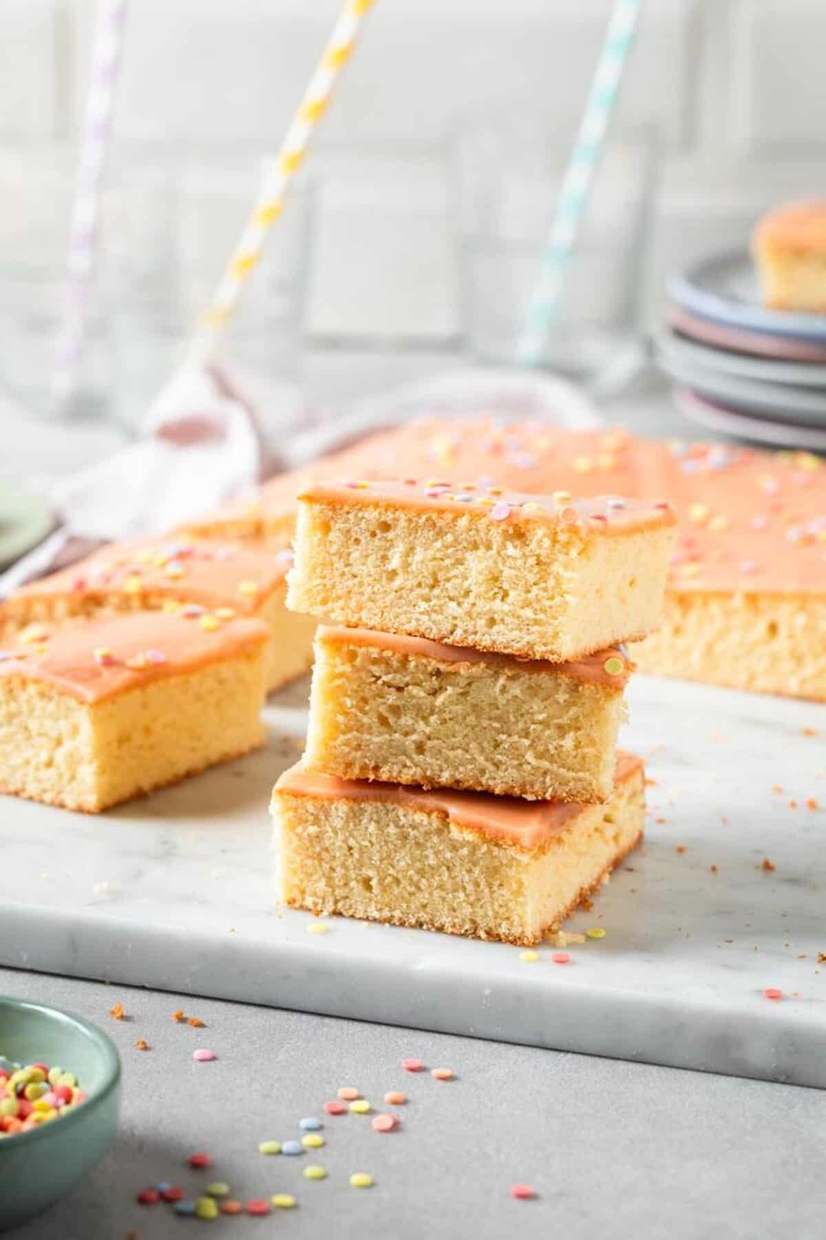 Flacher Blechkuchen mit Zuckerguss in Orange und Streuseln, mehrere Stücke gestapelt auf einem Teller – Foto: Anna Gieseler