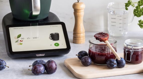 Ein Thermomix® TMt, davor liegen Pflaumen, auf einem Brett stehen Gläser mit Pflaumenmarmelade