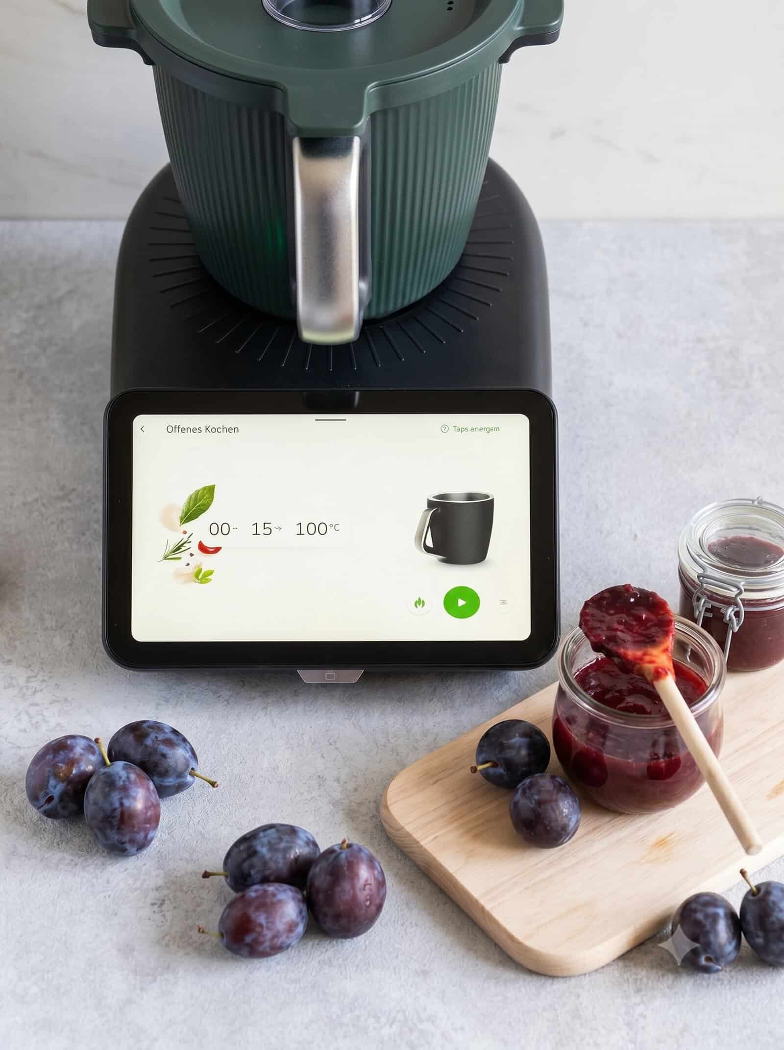 Ein Thermomix® TMt, davor liegen Pflaumen, auf einem Brett stehen Gläser mit Pflaumenmarmelade