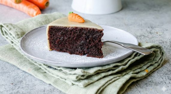 Ein Stück Schokoladenkuchen mit Marzipandecke und einer Karotte auf einem hellen Teller. Sehr saftig