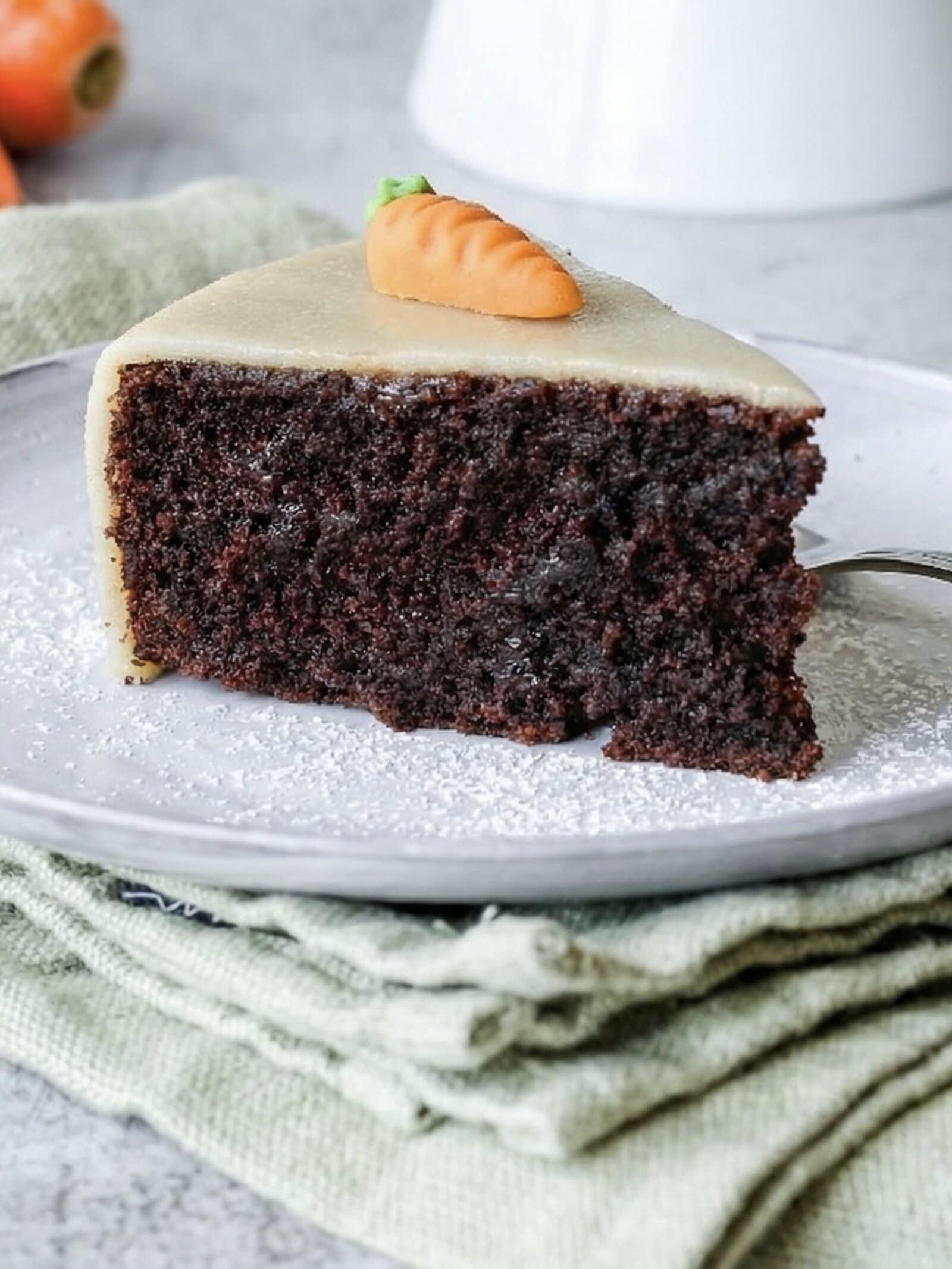 Ein Stück Schokoladenkuchen mit Marzipandecke und einer Karotte auf einem hellen Teller. Sehr saftig