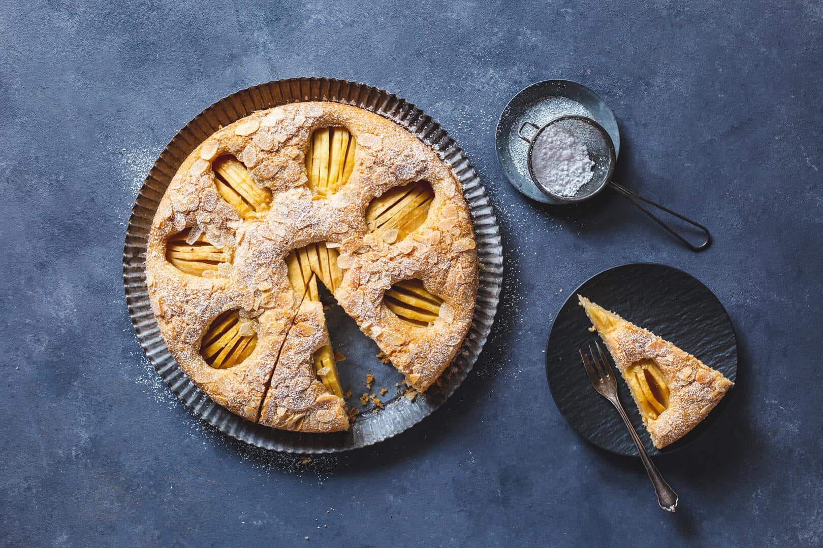Apfelkuchen mit versunkenen Äpfeln, angeschnitten auf grauem Untergrund