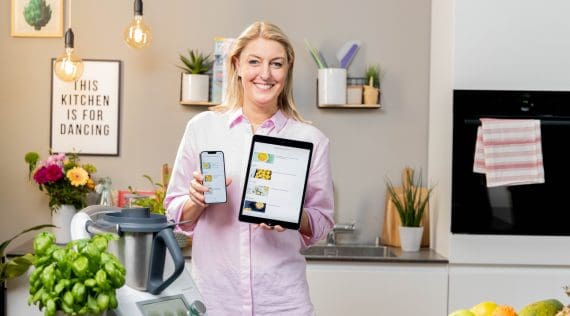 Eine blonde Frau steht in eienr Küche neben dem Thermomix® und hält ein Smartphone und ein Tablet hoch. Darauf der ZauberTopf Club
