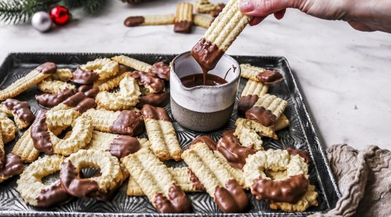 Ein Blech voll Spritzgebäck, eine Hand taucht ein Plätzchen in flüssige Schokolade – Foto: Sandra Leibinger