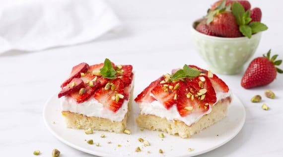 Zwei Stücke Erdbeerkuchen auf einem hellen Teller, im Hintergrund eine Schale mit Erdbeeren