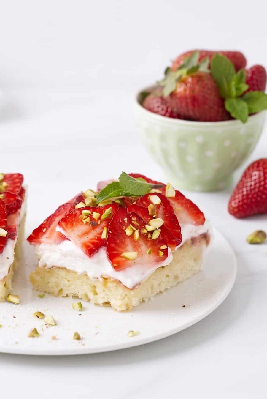 Zwei Stücke Erdbeerkuchen auf einem hellen Teller, im Hintergrund eine Schale mit Erdbeeren