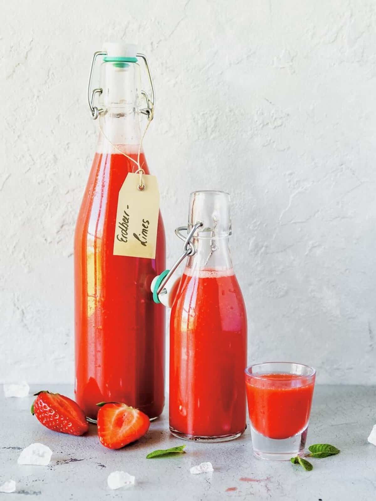 Zwei Flaschen mit rotem Erdbeer-Limes und ein Glas vor grauem Hintergrund – Foto: Désirée Peikert