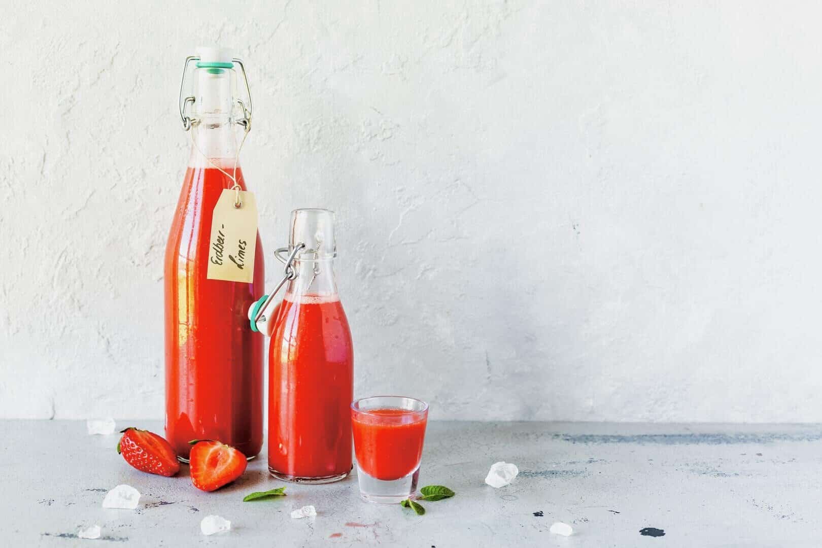 Zwei Flaschen mit rotem Erdbeer-Limes und ein Glas vor grauem Hintergrund – Foto: Désirée Peikert