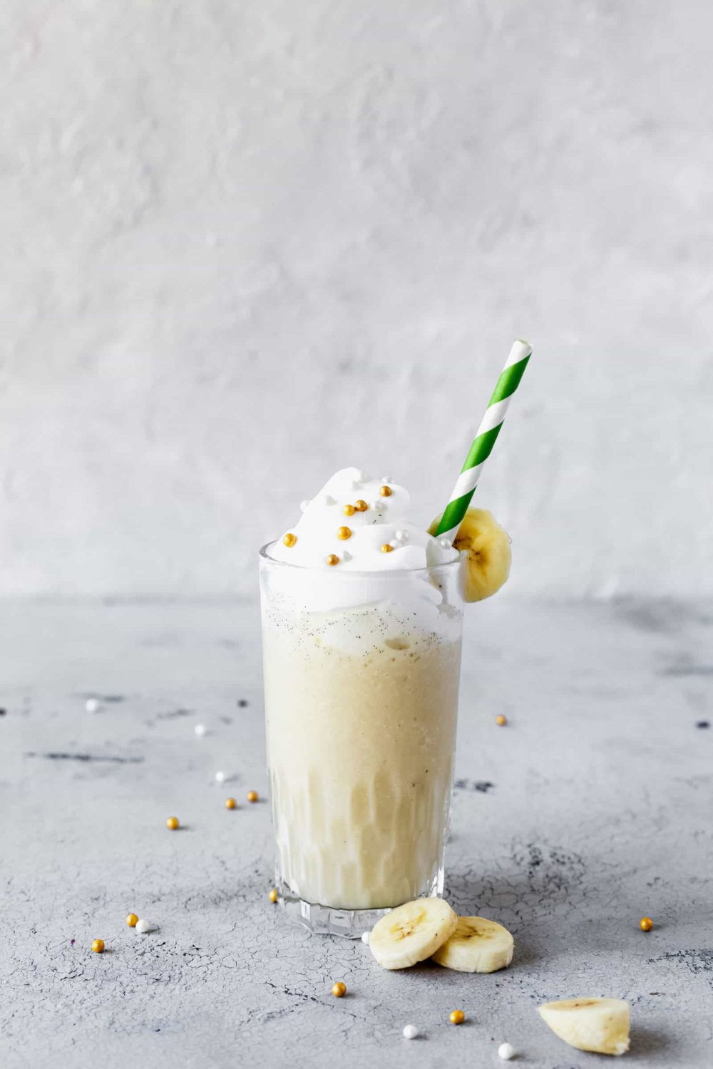 Ein Glas Bananen-Milchshake mit Sahne und Zuckerperlen dekoriert, darin ein grün-weiß gestreifter Strohhalm