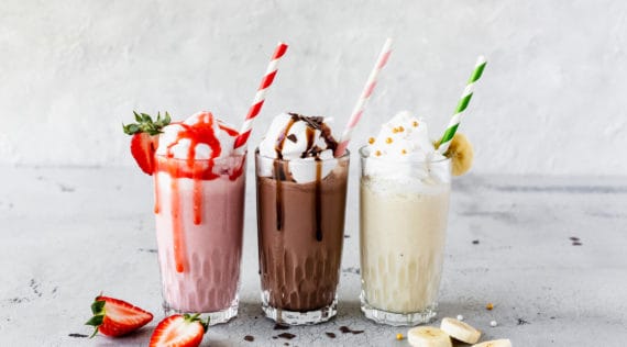 Drei Gläser mit Milchshakes: Erdbeer, Schokolade und Banane, jeweils mit Sahne und einem Strohhalm dekoriert