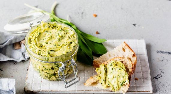 Cremige Butter mit Kräutern in einem Glas, daneben Bärlauch und ein Stück Brot mit der Butter beschmiert