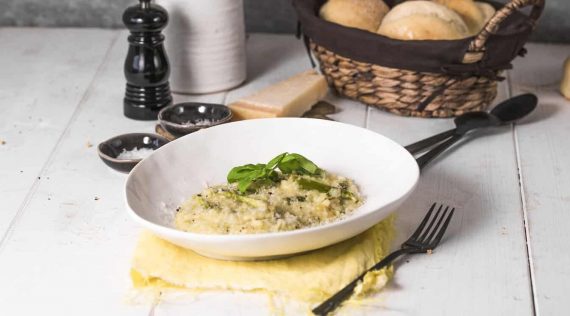Ein Teller mit Spargelrisotto auf einem weißen Holztisch, im Hintergrund Blumen und ein Korb mit Brötchen
