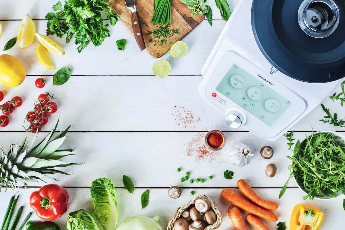 Buntes Gemüse und die Küchenmaschine Monsieur Cuisine auf einem weißen Holzuntergrund