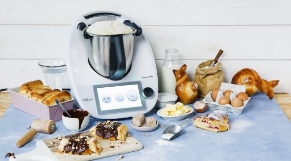 Ein Thermomix® mit Hefeteig im Topf, umgeben von Gebäck, Brot, Croissants.