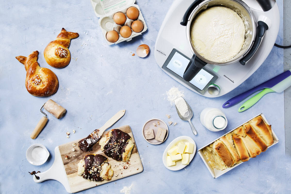 Ein Thermomix® mit Hefeteig im Mixtopf, umgeben von Eiern und Gebäck