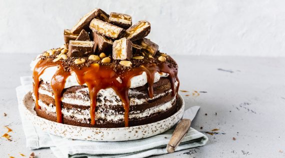 Snickers-Torte mit Karamellsoße und zerkleinerten Schokoriegeln auf einer weißen Platte