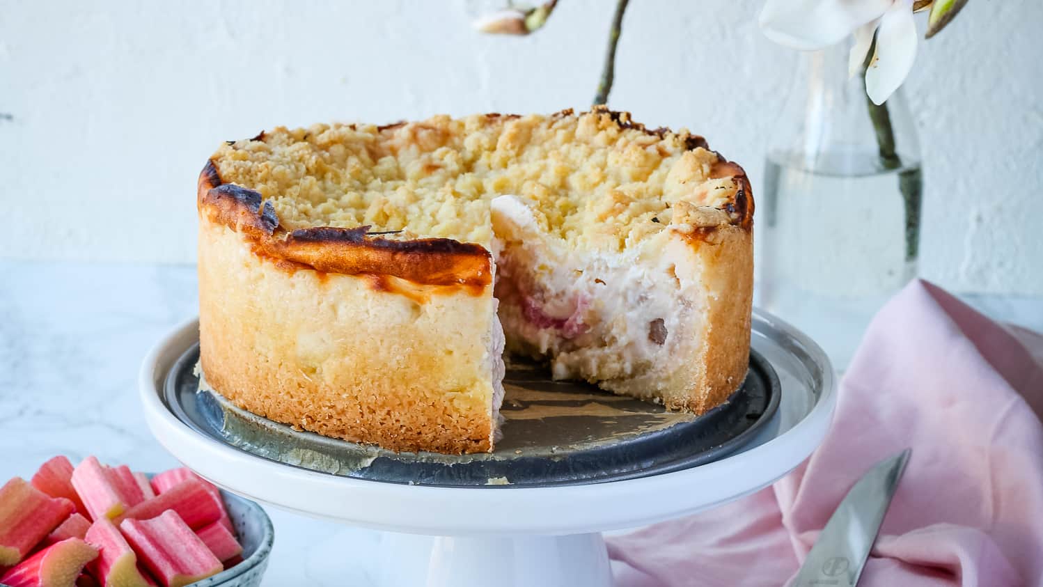 Rhabarber-Käsekuchen mit Streusel, angeschnitten auf einer weißen Tortenplatte