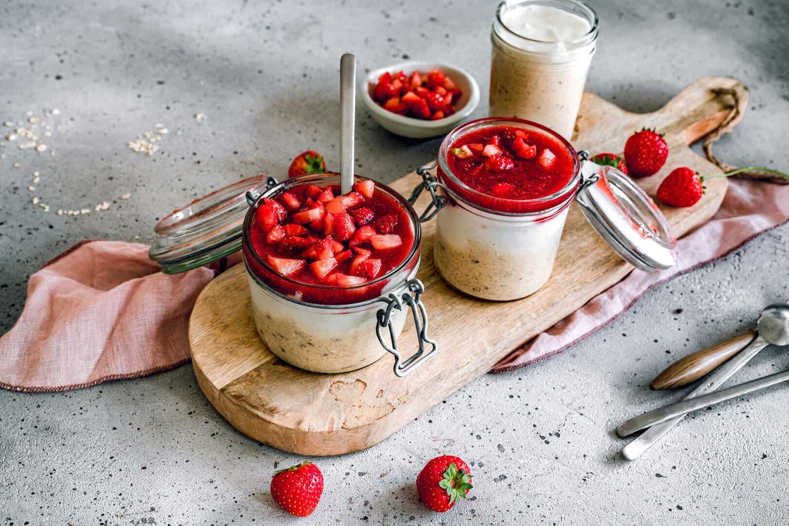 Zwei Gläser mit Haferflocken, Joghurt und einer Erdbeersoße auf einem Holzbrett