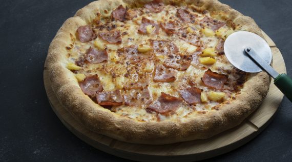 Pizza Hawaii mit Schinken und Ananas auf einem dunklen Untergrund mit Pizzaschneider