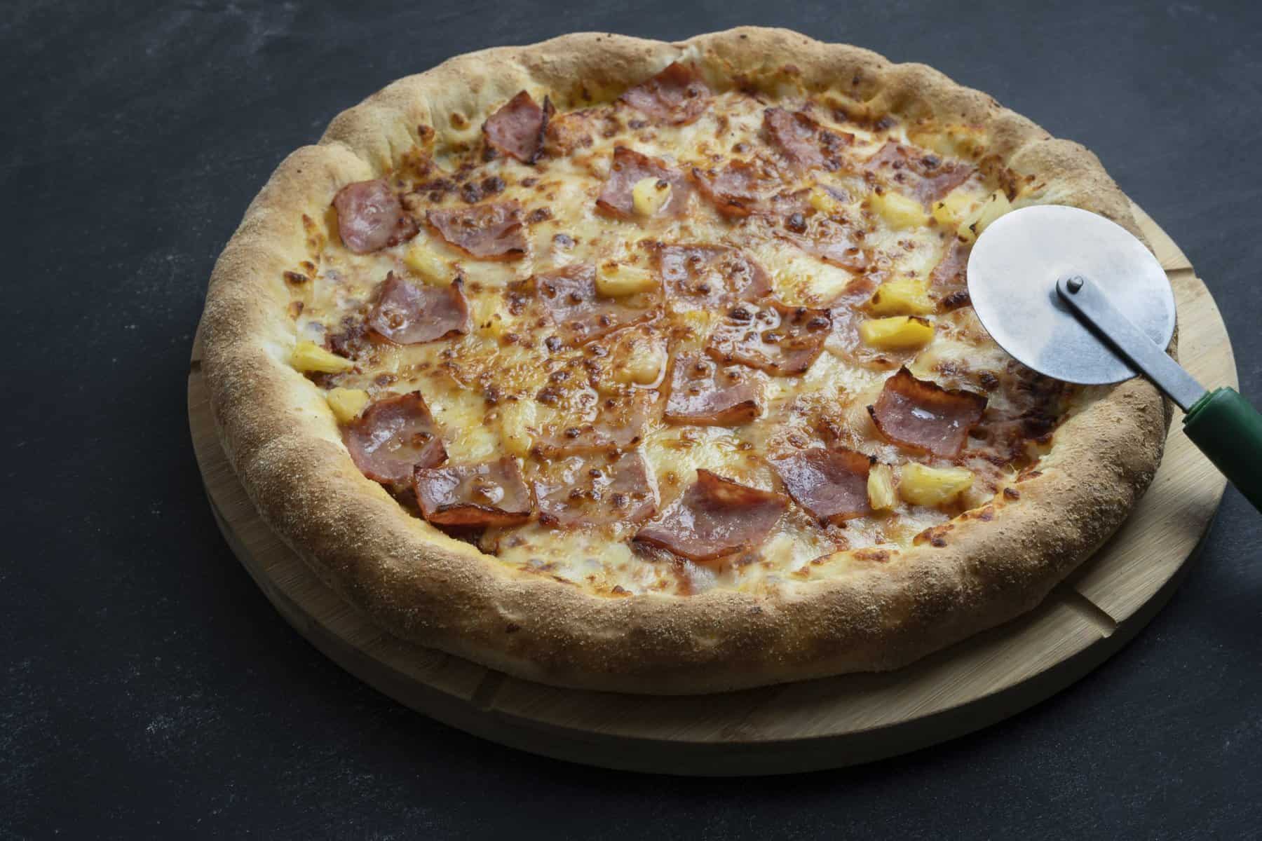 Pizza Hawaii mit Schinken und Ananas auf einem dunklen Untergrund mit Pizzaschneider