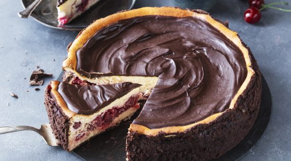 Schwarzwälder Käsetorte, angeschnitten auf einer dunlen Platte, im Hintergrund ein Glas und Kirschen