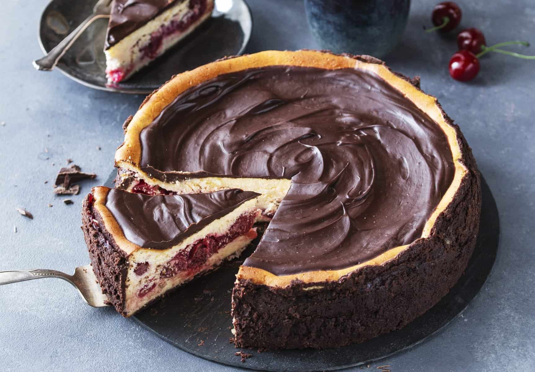 Schwarzwälder Käsetorte, angeschnitten auf einer dunlen Platte, im Hintergrund ein Glas und Kirschen