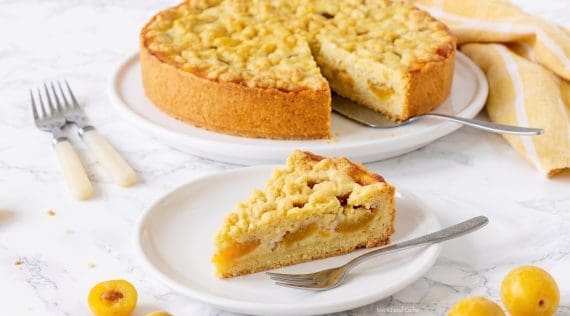 Ein Streuselkuchen mit Mirabellen darin auf einem weißen Teller, davor ein einzelnes Stück