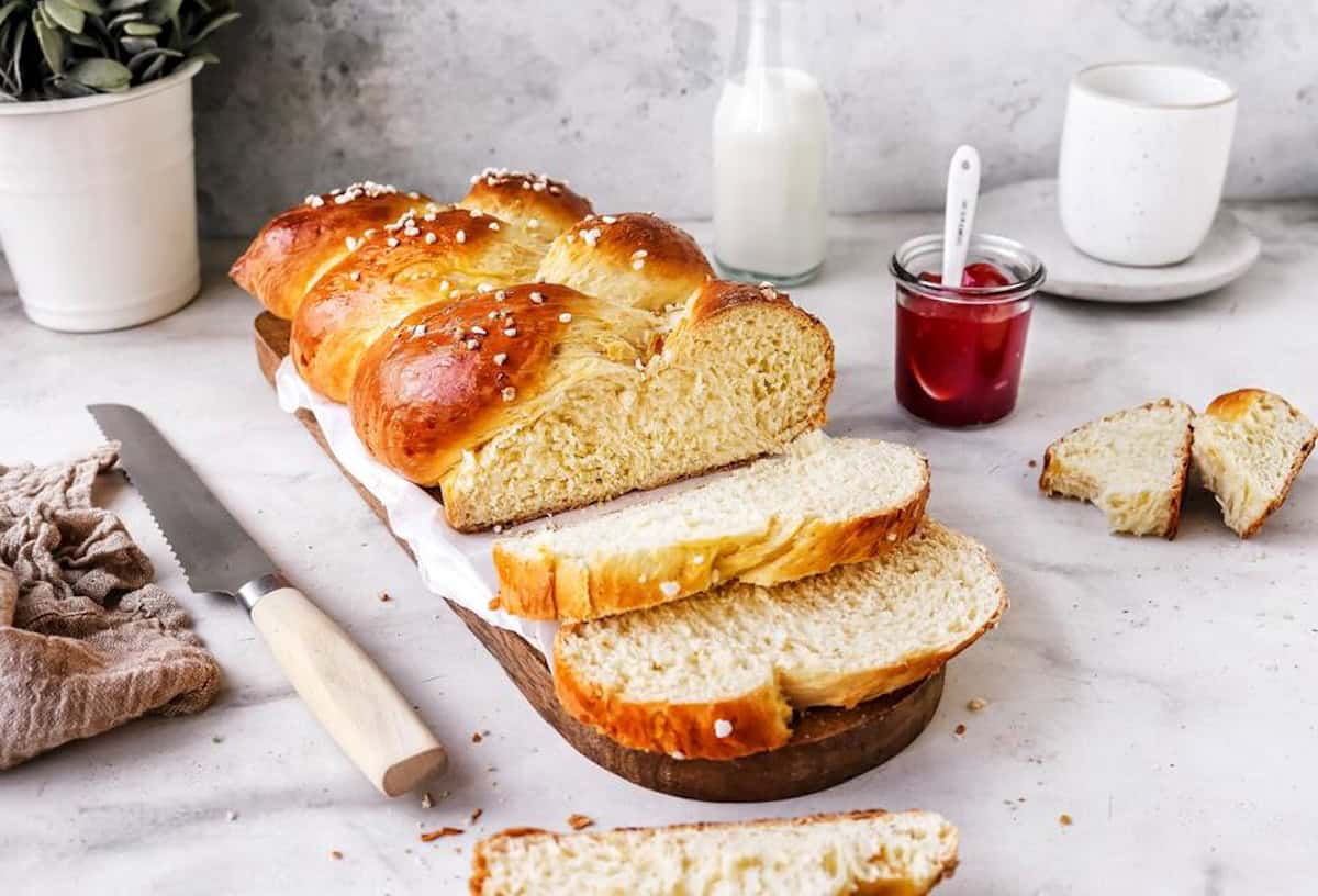 Saftiger Hefezopf, angeschnitten auf hellem Untergrund, daneben ein Glas Marmelade