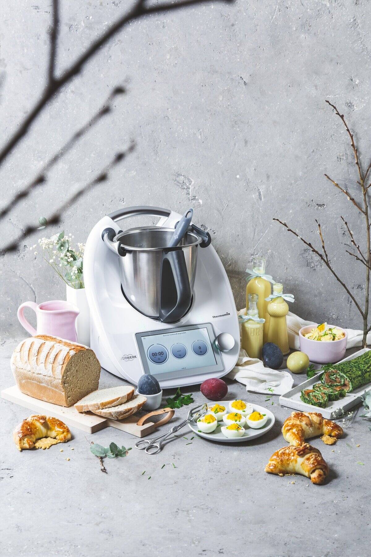 Ein weißer Thermomix®, umgeben von Croissants, Eierlikör, Brot, gefärbten Eiern