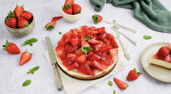 Ein Erdbeerkuchen auf hellem Untergrund, daneben Erdbeeren und Besteck