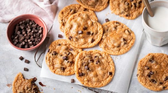 Mehrere Chocolate Chip Cookies auf weißem Backpapier, daneben Schokoladen-Stückchen in einer Schale