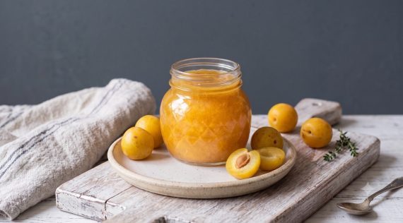 Ein Glas mit gelber Marmelade auf einem grauen Teller, daneben liegen Mirabellen