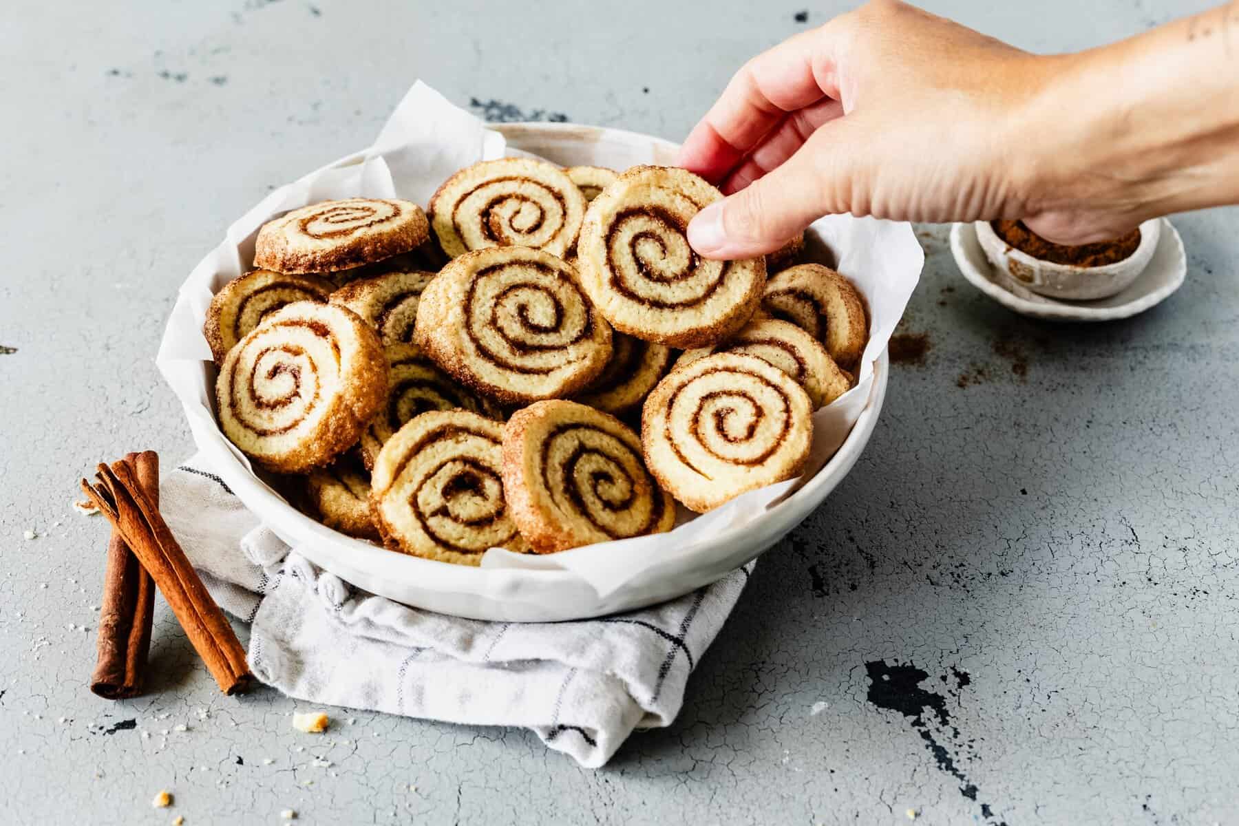 Eine weiße Schale mit runden Keksen, die aussehen wie Zimtschnecken. Eine Hand nimmt einen der Kekse – Foto: Désirée Peikert