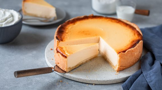 Ein angeschnittener Käsekuchen vor grauem Untergrund. Im Hintergrund Mehl und Milch
