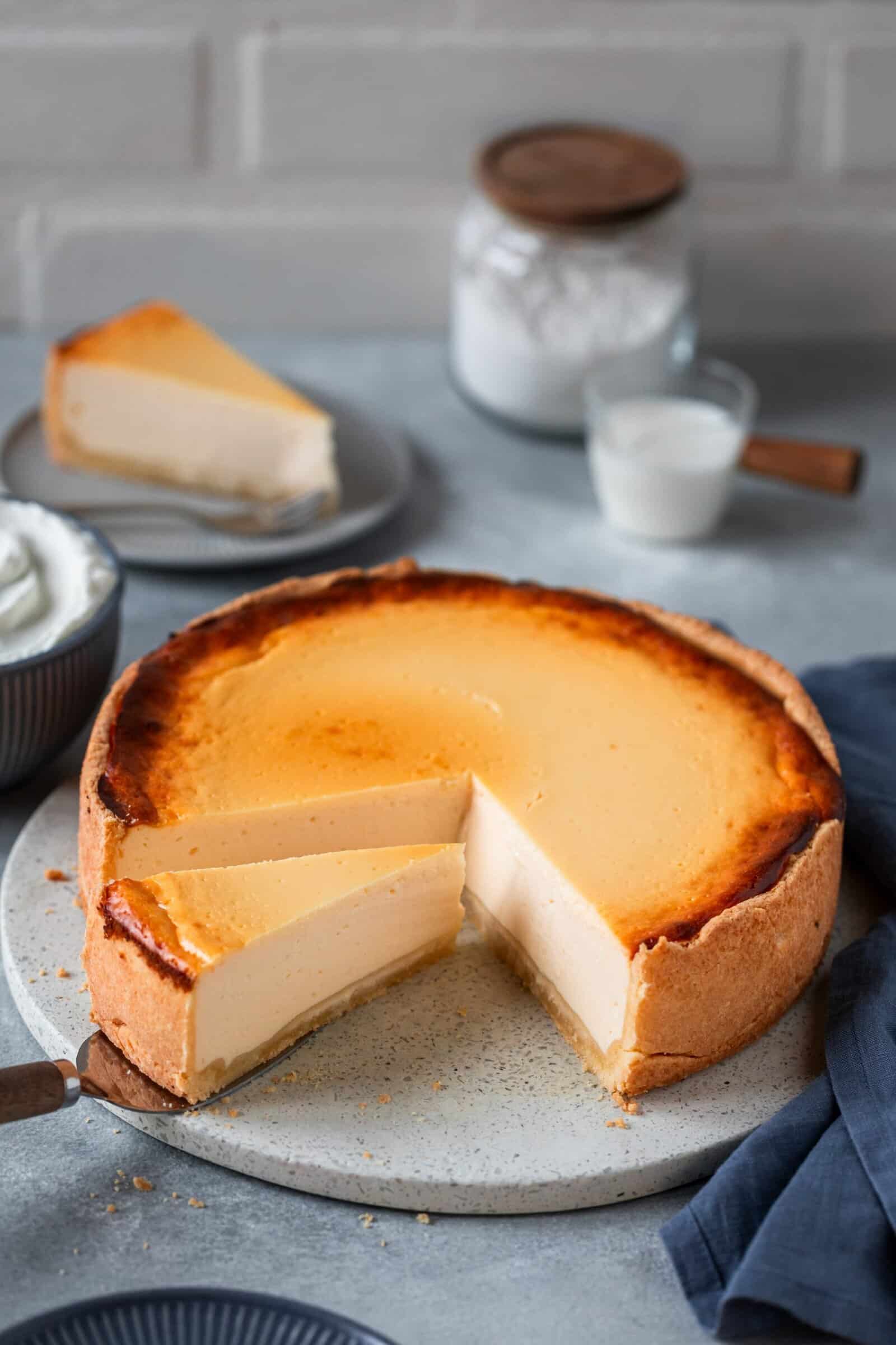 Ein angeschnittener Käsekuchen vor grauem Untergrund. Im Hintergrund Mehl und Milch