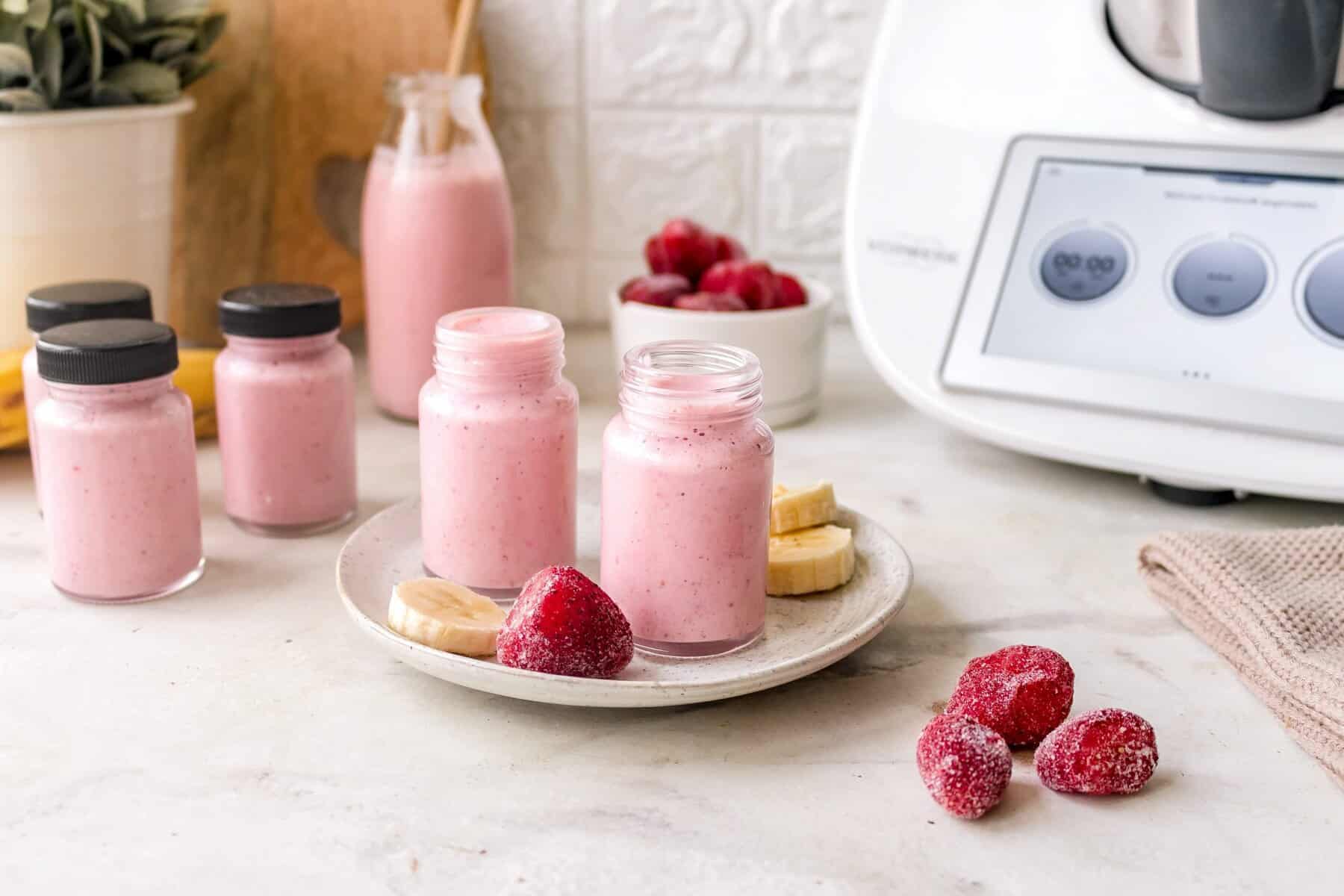 Vier kleine Flaschen mit rotem Joghurt-Getränk, daneben gefrorene Früchte, im Hintergrund ein Thermomix®