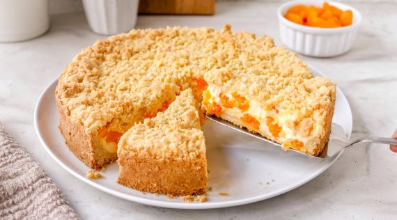 Ein Streuselkuchen mit Cremefüllung, die Mandarinen enthält, angeschnitten auf einem hellen Teller
