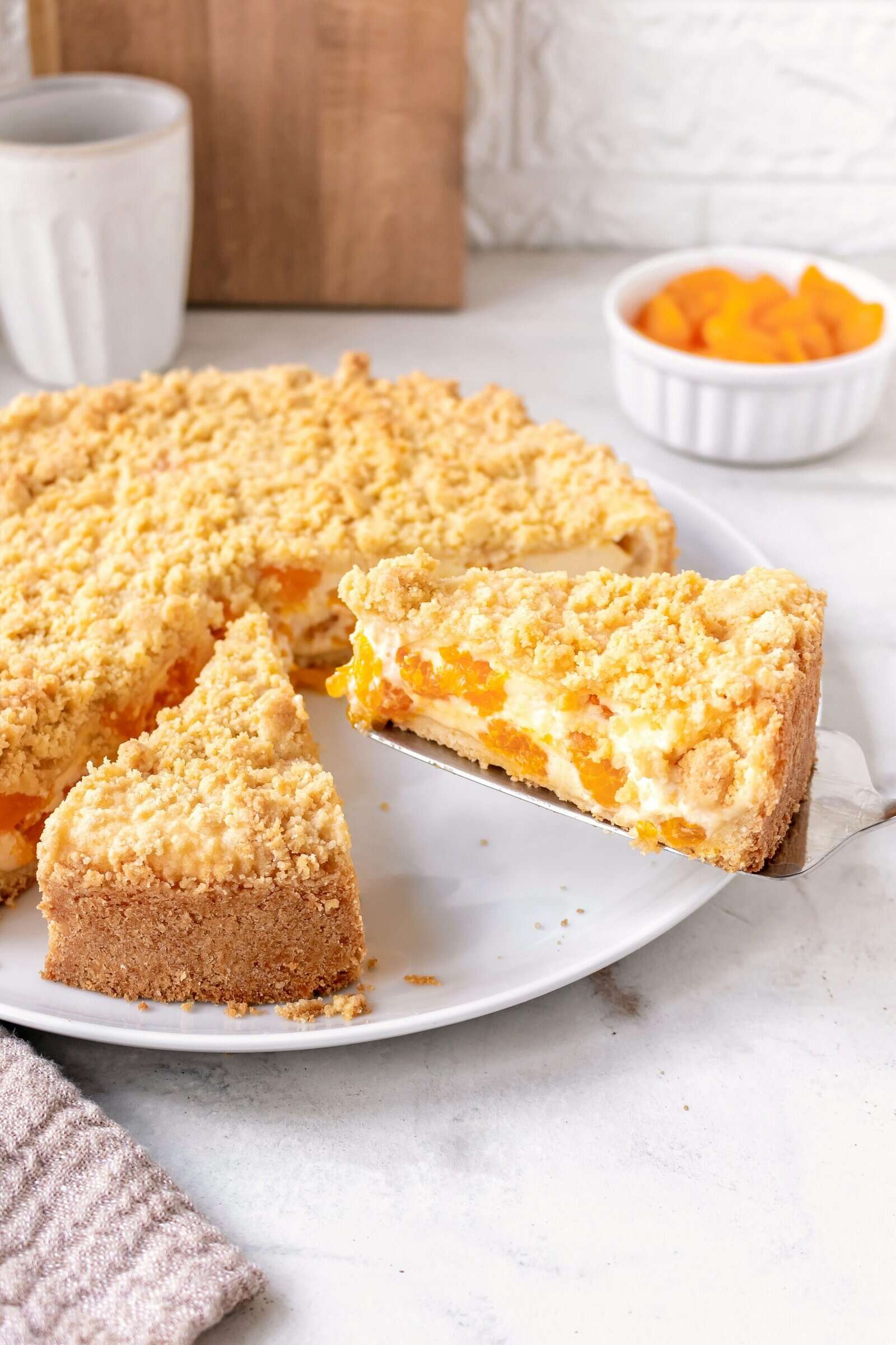 Ein Streuselkuchen mit Cremefüllung, die Mandarinen enthält, angeschnitten auf einem hellen Teller